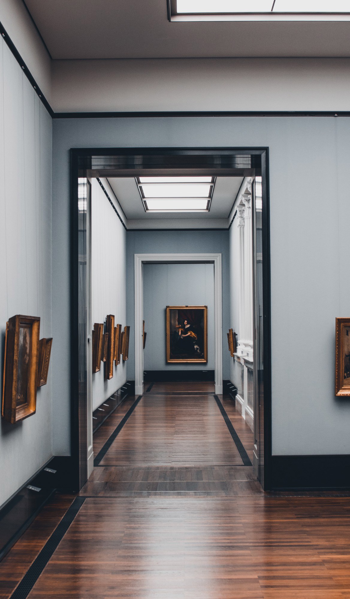 Doorkijk door meerdere museumzalen met lijstschilderijen aan lichtblauwe wanden, een glanzende houten vloer en een verlicht portret in de verte.