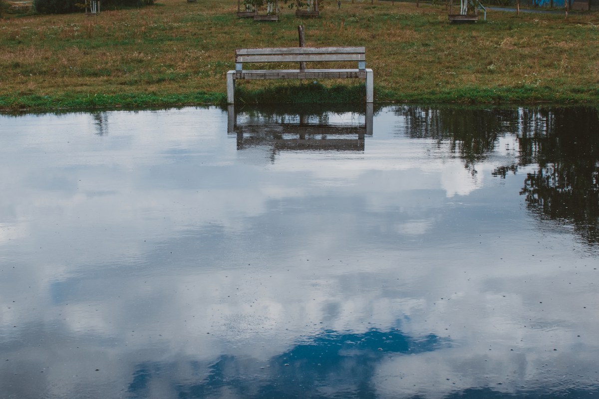 Een houten bankje staat half onder water in een ondergelopen grasveld, waarin de bewolkte lucht zich weerspiegelt.
