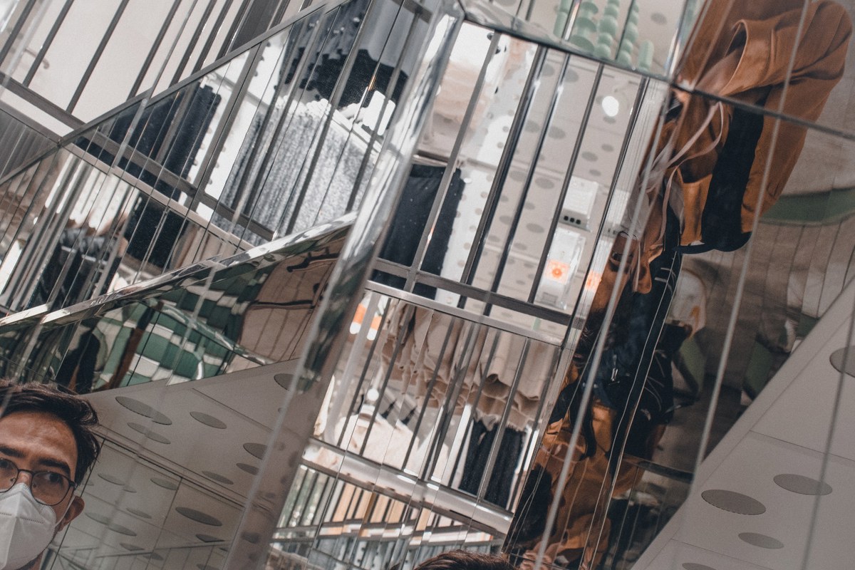 Spiegelend metalen kunstwerk met verticale lijnen weerkaatst een gefragmenteerd stadsbeeld, terwijl linksonder een man met mondkapje en bril zichtbaar is.