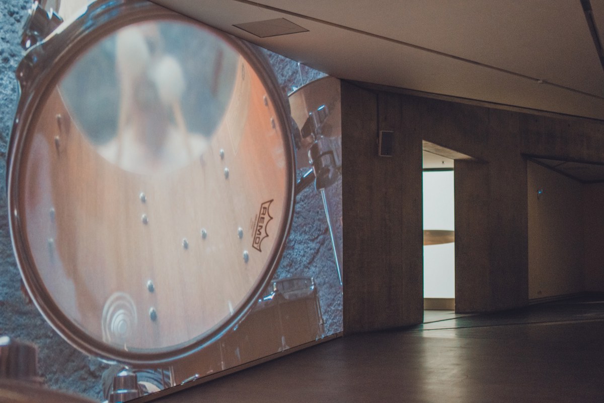 Een grote drum met Remo-vel domineert de voorgrond in een schemerige zaal met houten wanden en een verlichte doorgang op de achtergrond.