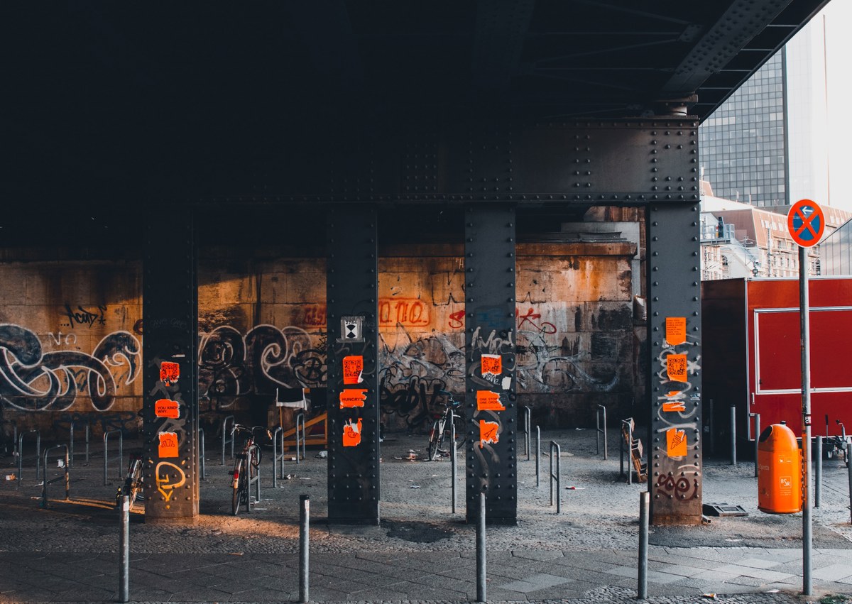 Onder een stalen spoorbrug staan zwarte pilaren voor met graffiti bedekte muren, met oranje posters en verdwijnende paaltjes in een verlaten stedelijke ruimte.