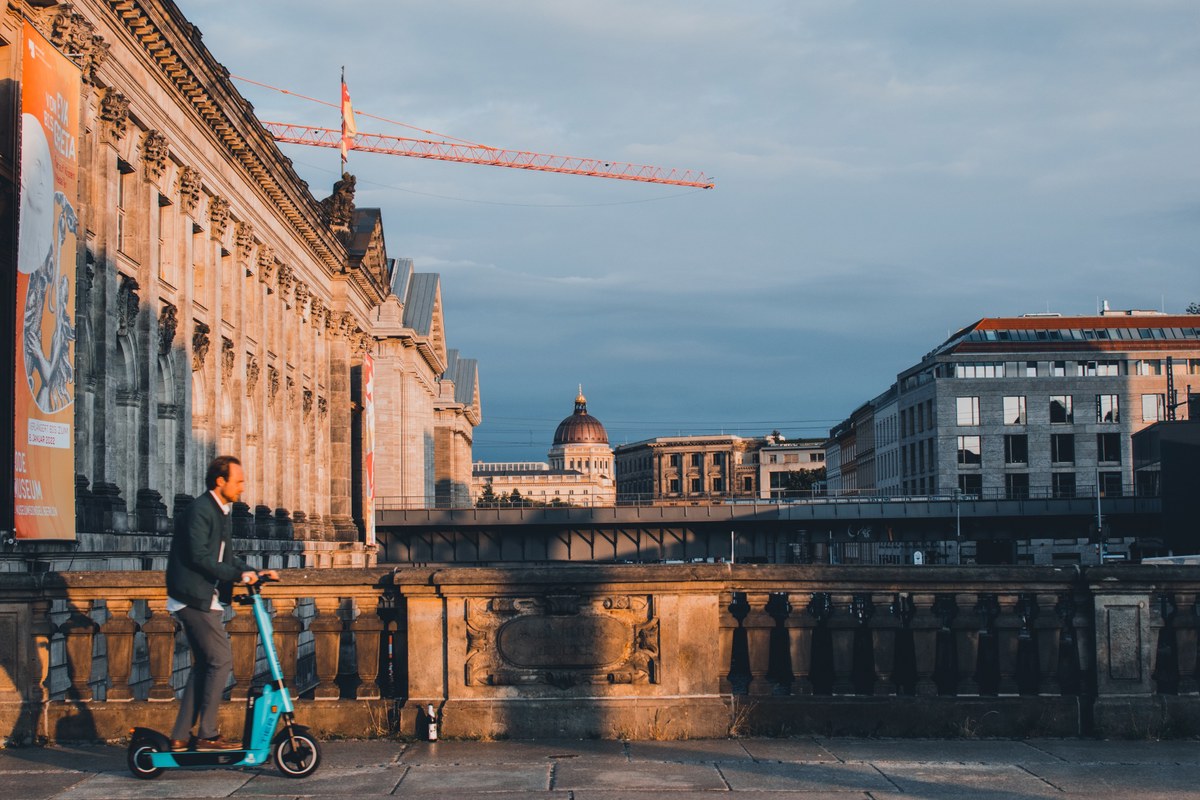 Een man op een turquoise elektrische step rijdt langs een historisch gebouw met een balustrade, terwijl op de achtergrond een bouwkraan en koepel opdoemen.