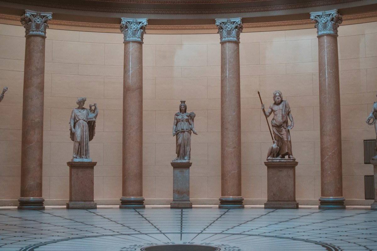 Klassieke marmeren beelden staan op sokkels tussen hoge Korinthische zuilen in een halfronde museumzaal met een geometrisch betegelde vloer.