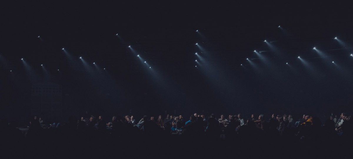 Rijen witte spotlights schijnen diagonaal door de duisternis boven een silhouet van een publiek tijdens de audiovisuele show Sora by Nonotak.