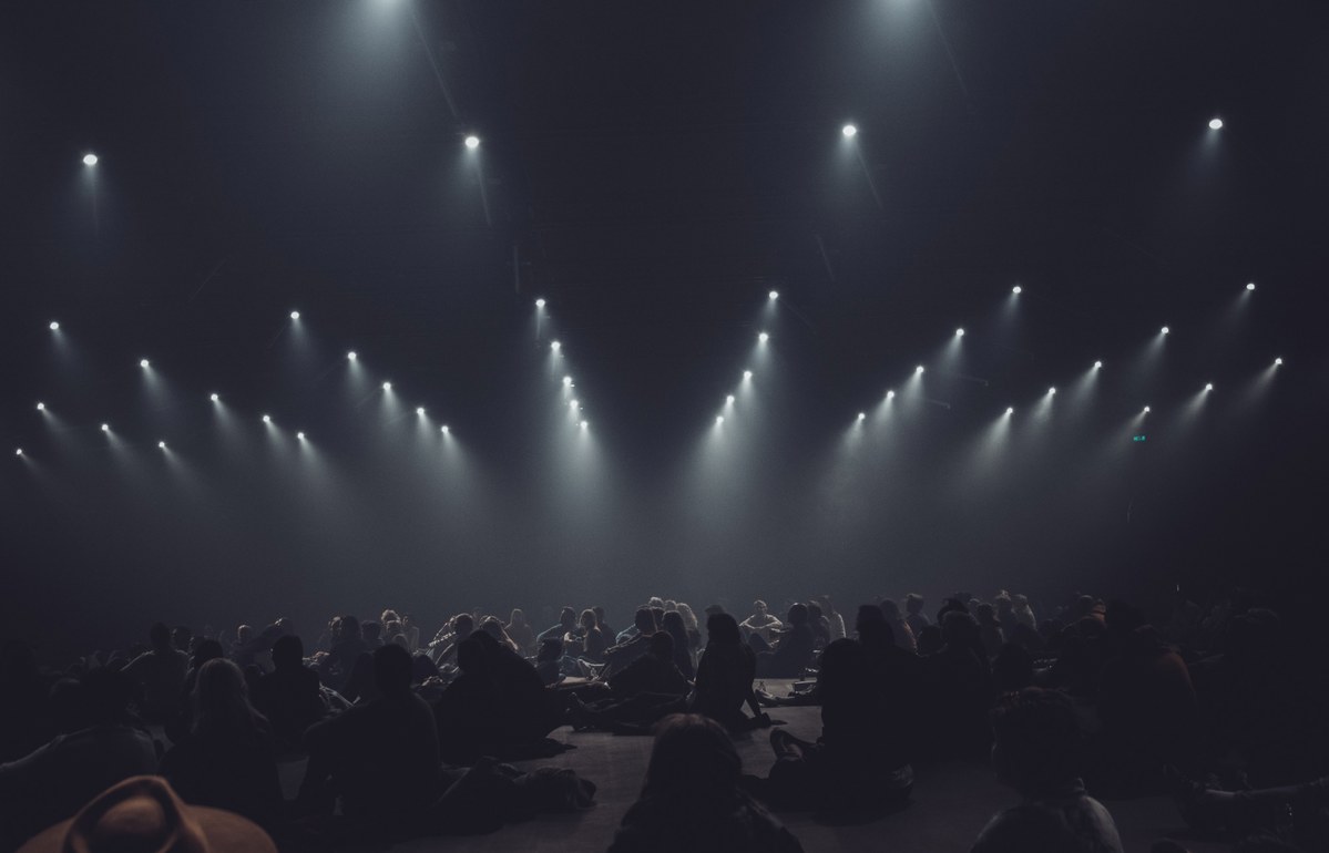 Silhouetten van een zittend publiek in een donkere zaal, verlicht door rijen felle spots die V-vormig neerstralen door de mist.