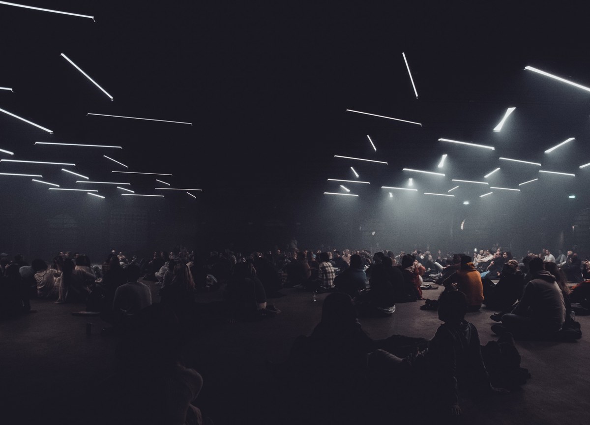 Een groot publiek zit in het donker op de vloer onder een plafond vol zwevende lichtstrepen tijdens een lichtinstallatie.