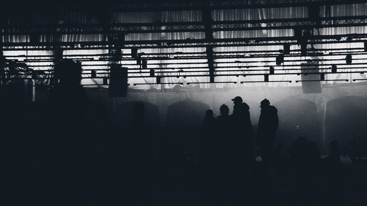 Silhouetten van mensen staan in een mistige, industriële hal met een verlicht plafond vol lichtbalken, in zwart-wit vastgelegd.