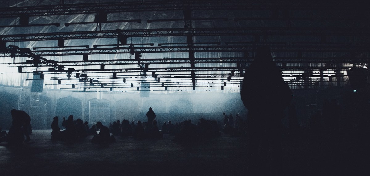 Silhouetten van bezoekers zitten en staan in een mistige, industriële hal met stalen dakspanten, terwijl blauwig licht door het rook wordt uitgelicht.