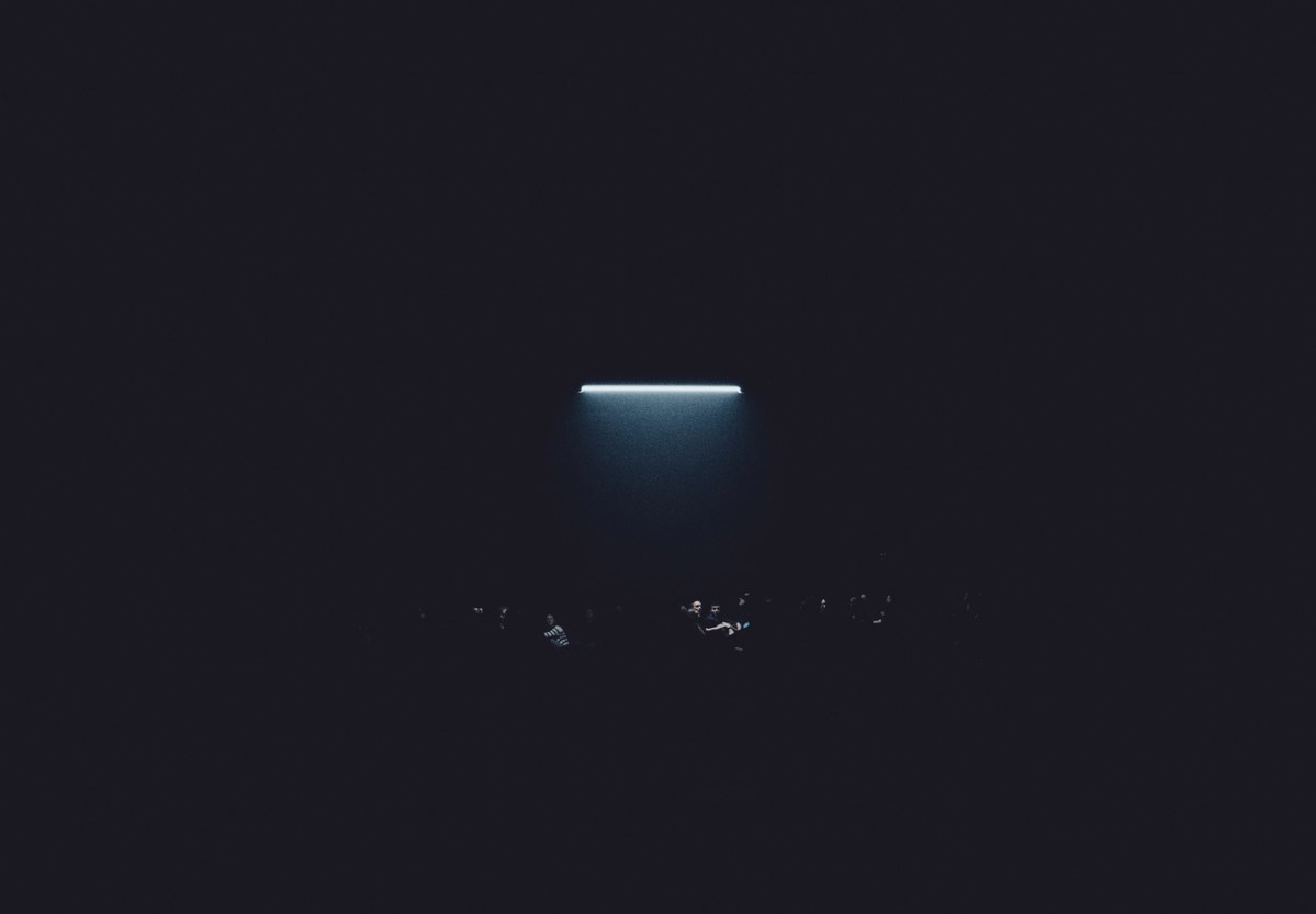 Een fel verlichte horizontale lichtbalk gloeit boven een donkere ruimte waarin de vage silhouetten van toeschouwers nauwelijks zichtbaar zijn.