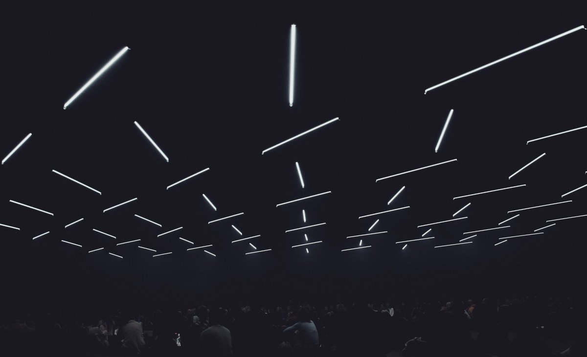 Talrijke felle lichtbuizen zweven op verschillende hoogtes onder een donker plafond boven de silhouetten van een publiek in een zaal.