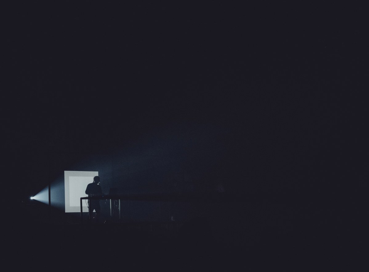 Silhouet van een persoon achter een tafel in een grotendeels donkere ruimte, verlicht door een smalle lichtbundel en een verlicht vlak links.