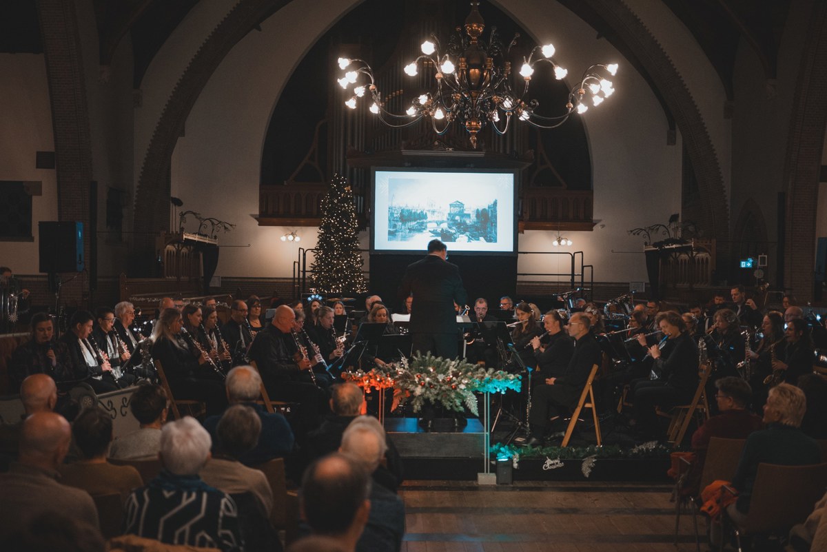Een dirigent leidt een orkest tijdens een kerstconcert in een sfeervol verlichte kerk, met een kerstboom, projectiescherm en aandachtig publiek.