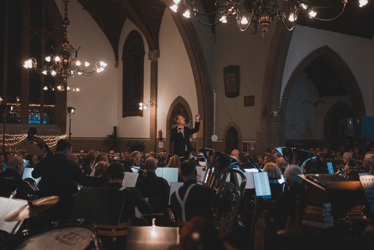 Een dirigent leidt met opgeheven armen een orkest tijdens een kerstconcert in een sfeervol verlichte kerk met kroonluchters en bogen.