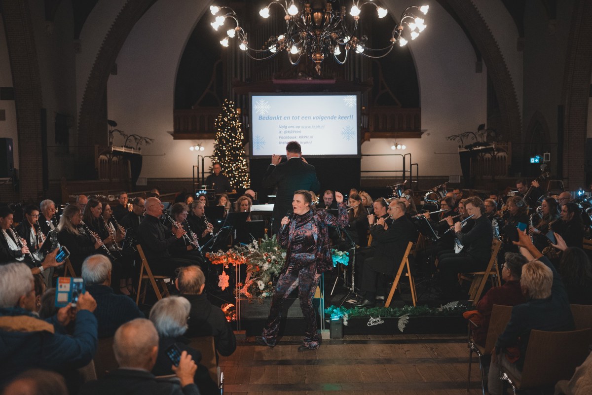 Een dirigent leidt een orkest in een sfeervol verlichte kerk tijdens een kerstconcert, terwijl het publiek toekijkt onder een grote kroonluchter.