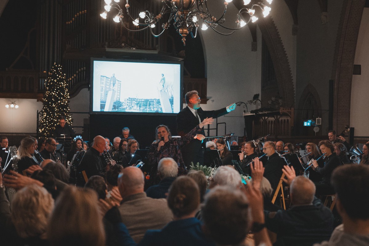Een dirigent leidt met gespreide armen een orkest tijdens een kerstconcert in een kerk, terwijl publiek op de voorgrond meeklapt onder sfeervolle kroonluchters.