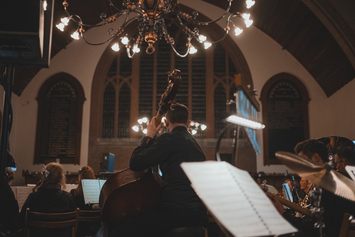Een contrabassist speelt met zijn rug naar de camera in een sfeervol verlichte kerk, omringd door orkestleden en muziekstandaards onder een grote kroonluchter.