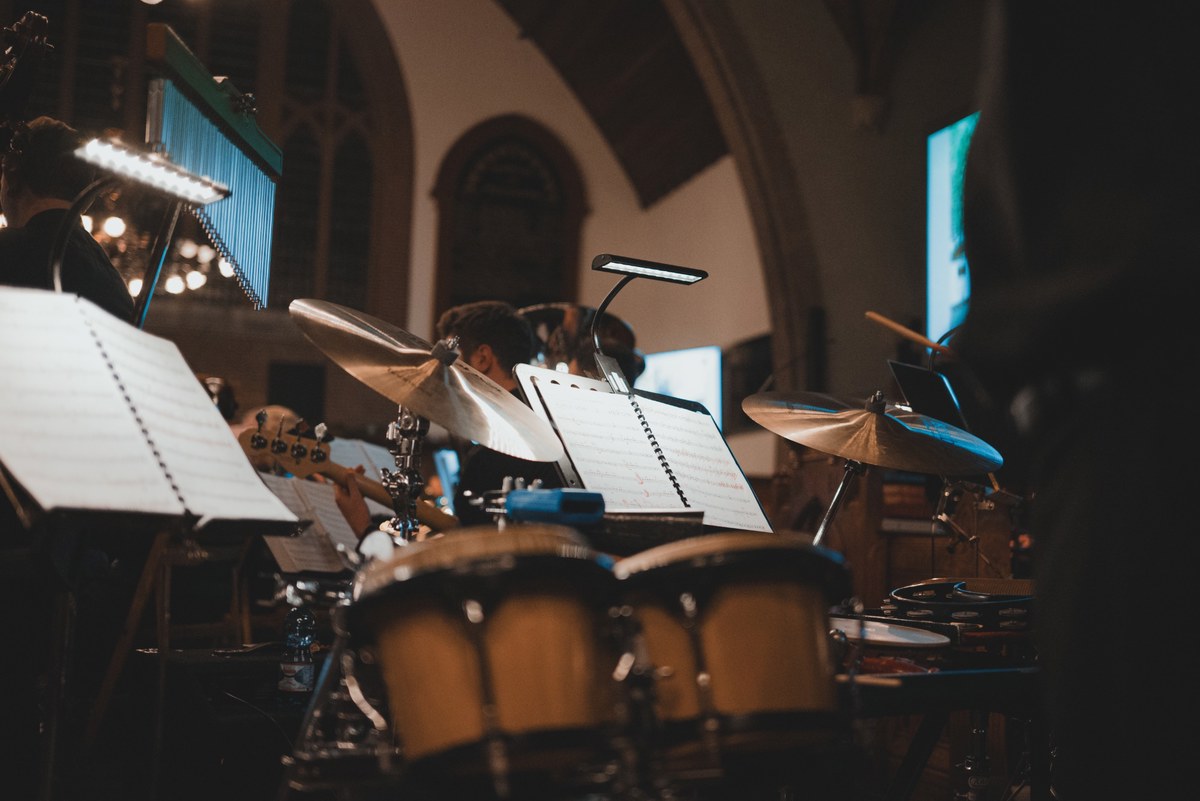 Muzikanten met bladmuziek op standaards spelen tijdens een kerstconcert in een kerk, met bekkens, bongo's en verlichte lessenaars op de voorgrond.