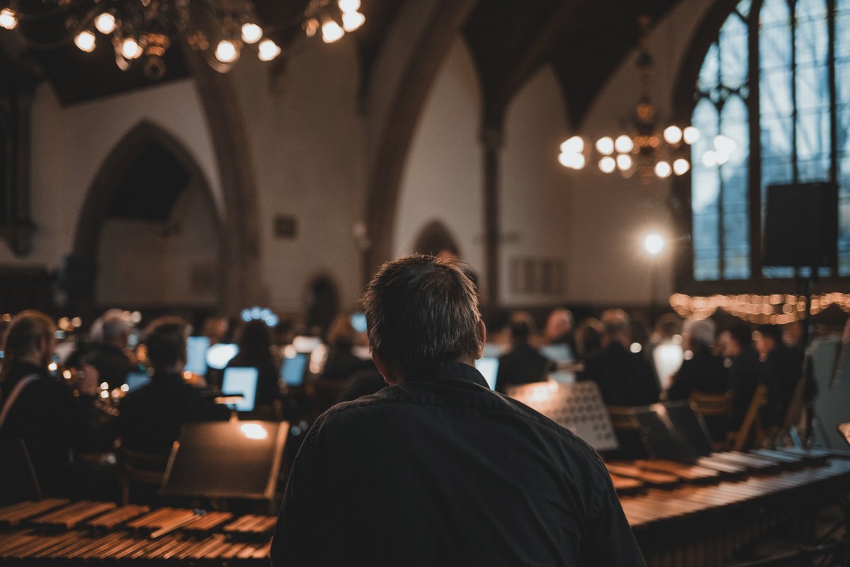 Van achteren gefotografeerde man kijkt naar een verlichte bijeenkomst in een sfeervolle kerk met hoge gotische ramen en warme lampjes.