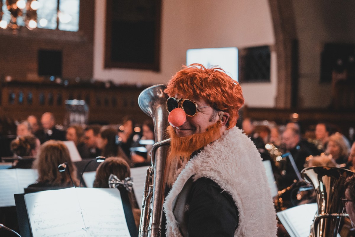 Muzikant met rode krullen, volle baard, ronde zonnebril en clownsneus bespeelt een tuba te midden van een orkest in een kerk.