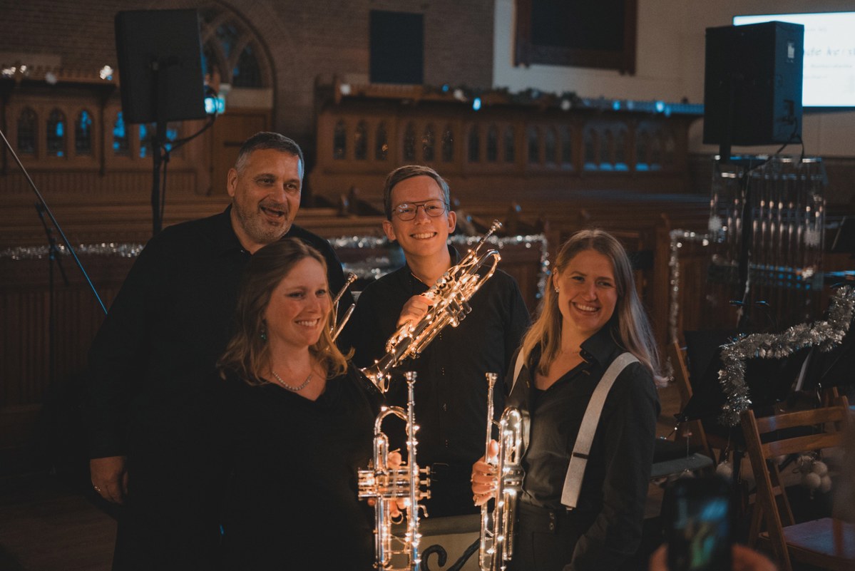 Vier lachende muzikanten poseren met glanzende trompetten in een sfeervol verlichte kerk tijdens een kerstconcert van KRPH.