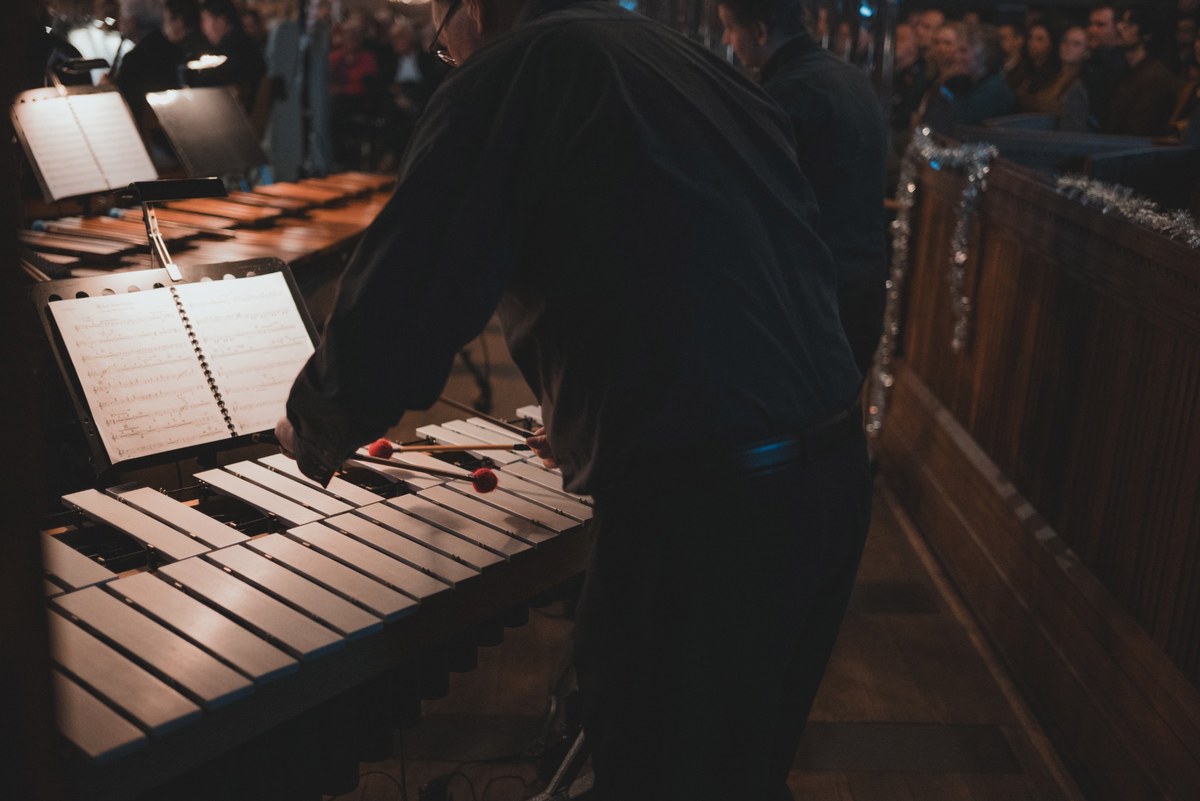 Een percussionist speelt met mallets op een marimba, met bladmuziek op de standaard en een aandachtig publiek op de achtergrond tijdens een kerstconcert.
