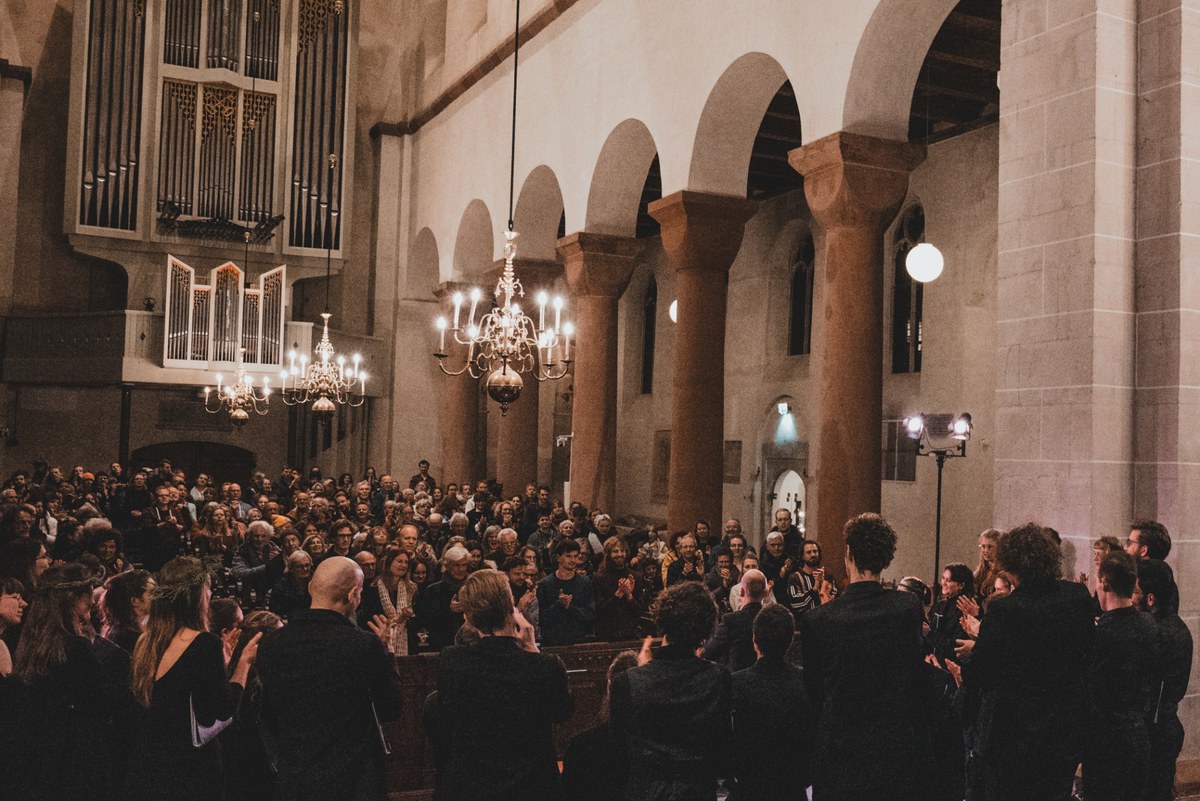 Publiek en koorleden applaudisseren in een sfeervol verlichte kerk met romaanse bogen, een groot orgel en hangende kroonluchters.
