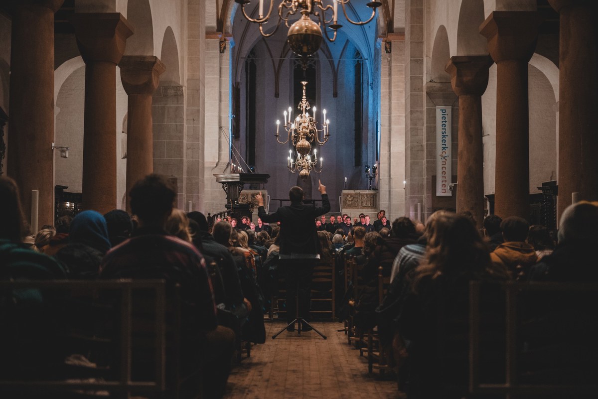 Een dirigent leidt vanaf een katheder een koor in een sfeervol verlichte kerk, terwijl het publiek in de banken toekijkt onder een gouden kroonluchter.