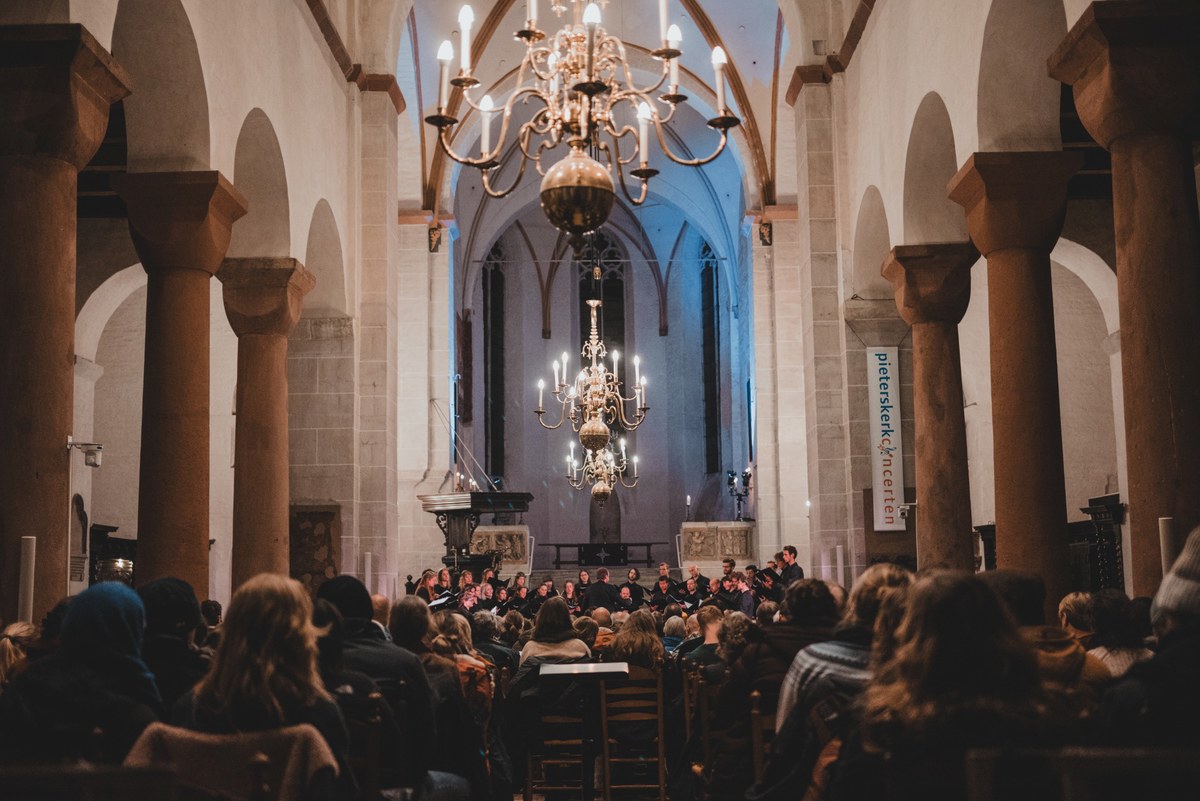 Publiek zit op houten stoelen in een sfeervol verlichte kerk terwijl een koor onder grote kroonluchters voor het altaar optreedt.