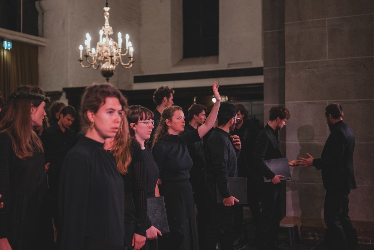 Koorleden in het zwart zingen in een kerk met kroonluchter, terwijl een dirigent hen met opgeheven hand aanstuurt tijdens een optreden.