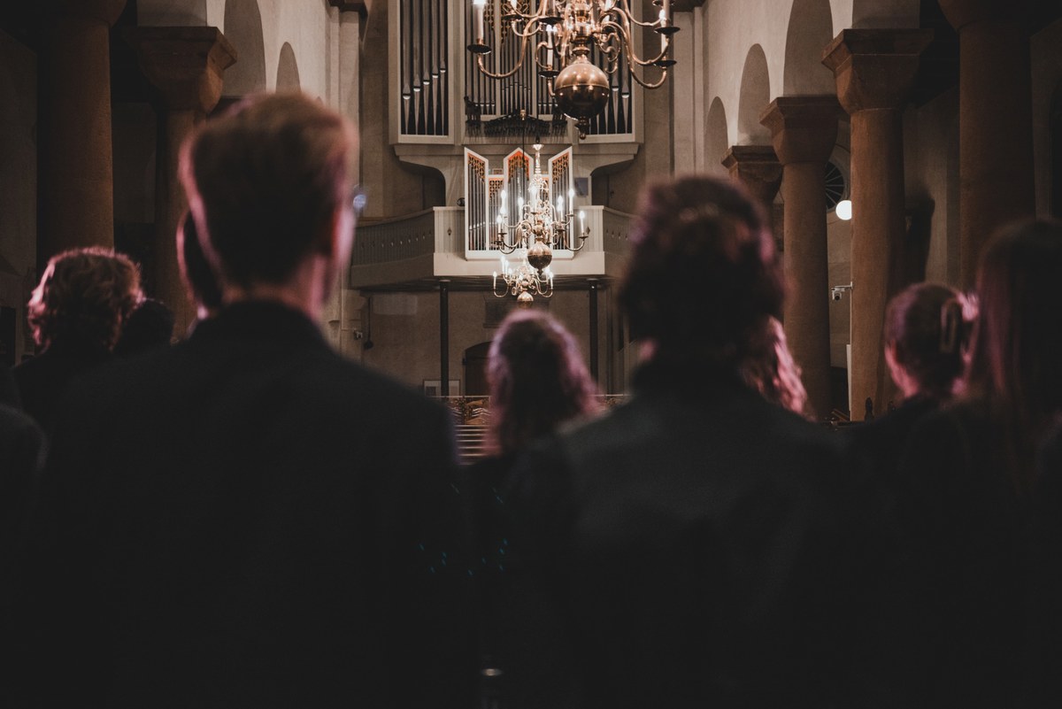 Publiek van achteren gezien in een sfeervol verlichte kerk kijkt naar een verlichte kroonluchter en orgel tijdens een concert van Kamerkoor JIP.