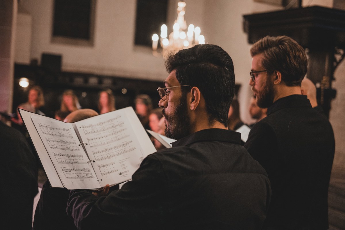 Twee koorzangers houden een muziekmap vast en lezen de bladmuziek tijdens een concert in een kerk, met een verlichte kroonluchter op de achtergrond.