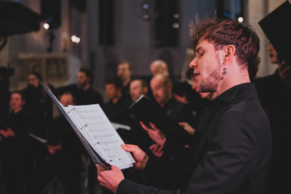 Een jonge zanger van Kamerkoor JIP zingt met bladmuziek in de hand, terwijl achter hem in een kerk overige koorleden in het donker meezingen.