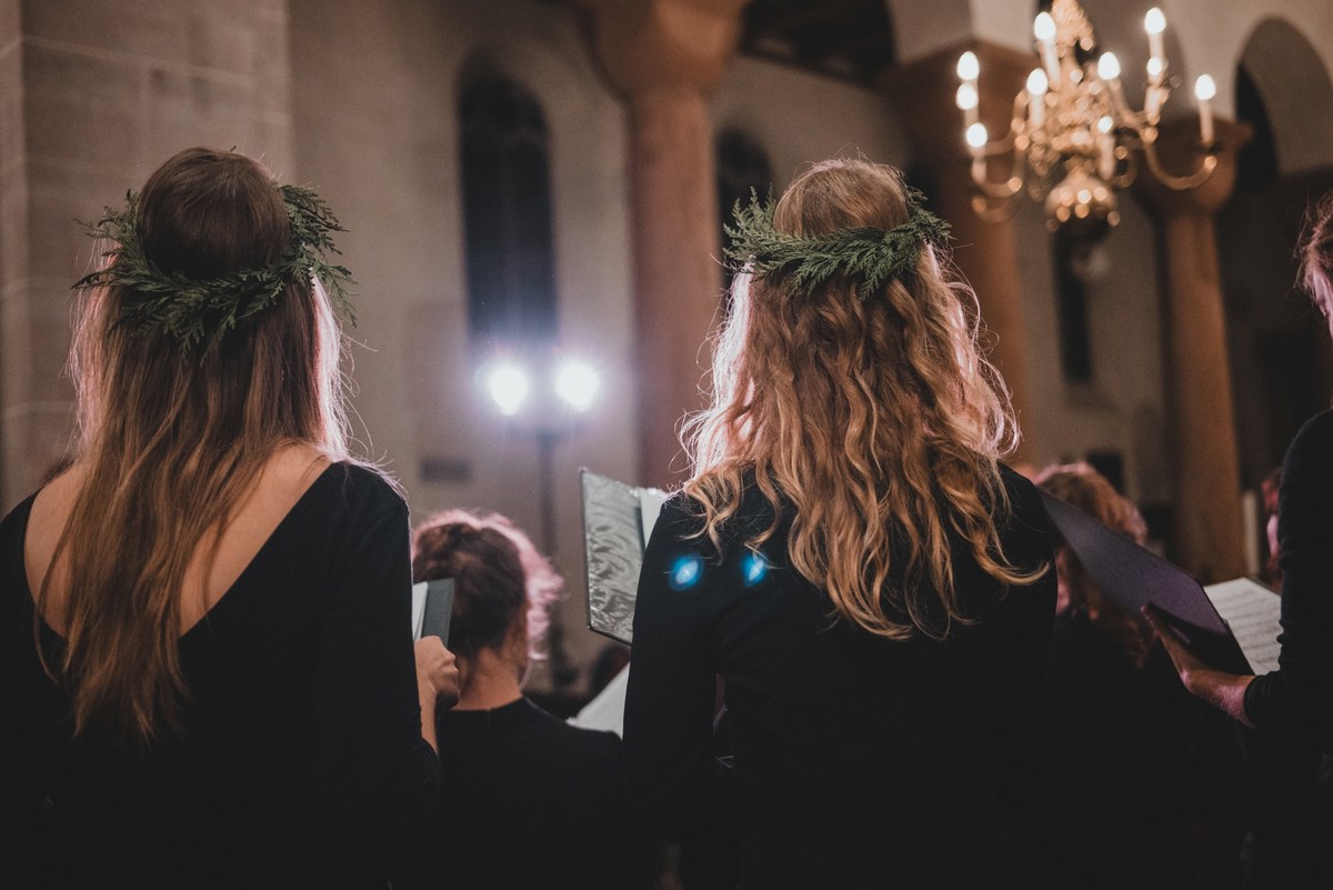 Twee vrouwen met groene bladerkransen in het haar staan van achteren gezien te zingen uit hun bladmuziek in een sfeervol verlichte kerk.