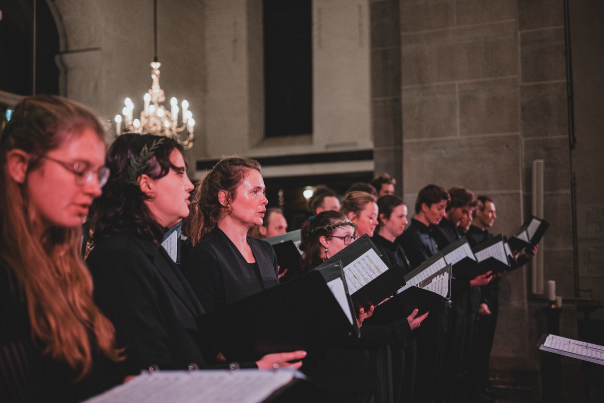Leden van Kamerkoor JIP zingen in het zwart gekleed uit hun bladmuziek in een kerk, met een kroonluchter en stenen muren op de achtergrond.