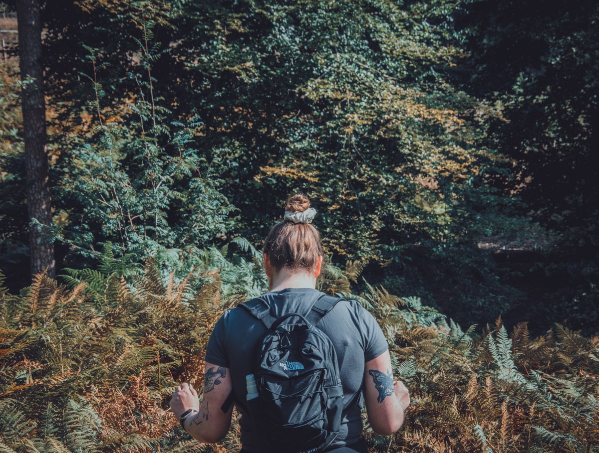 Van achteren gezien wandelt een persoon met haarknot, tatoeages en zwarte rugzak door dichte varens en groen bos op de Veluwe.