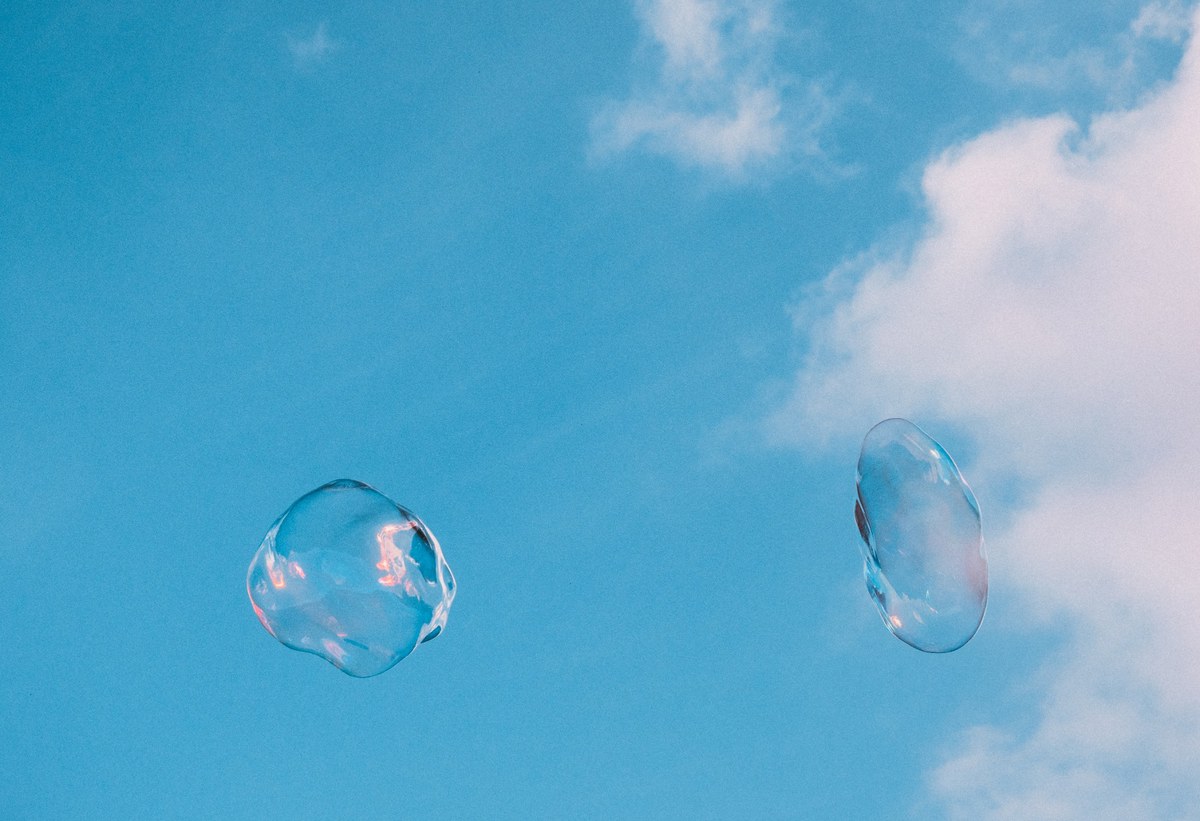 Twee grote zeepbellen zweven tegen een strakblauwe lucht met witte wolken, hun oppervlak weerspiegelt zachte roze en lila tinten.