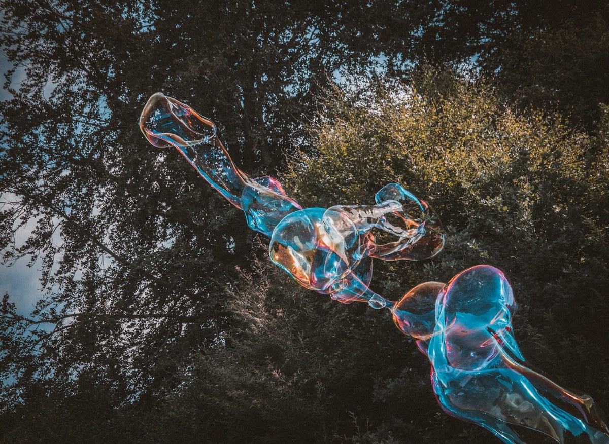 Een langgerekte reuzenzeepbel met blauwe en oranje glinsteringen zweeft tegen een achtergrond van donkere boomtoppen en bewolkte lucht.