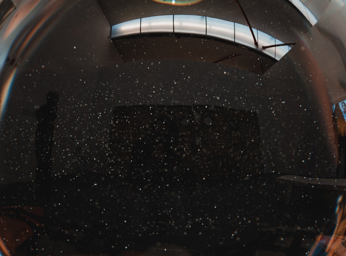 Silhouet van een persoon staand voor een donkere wand in een schemerige tentoonstellingsruimte, gezien door een fisheye-lens met stofdeeltjes in beeld.