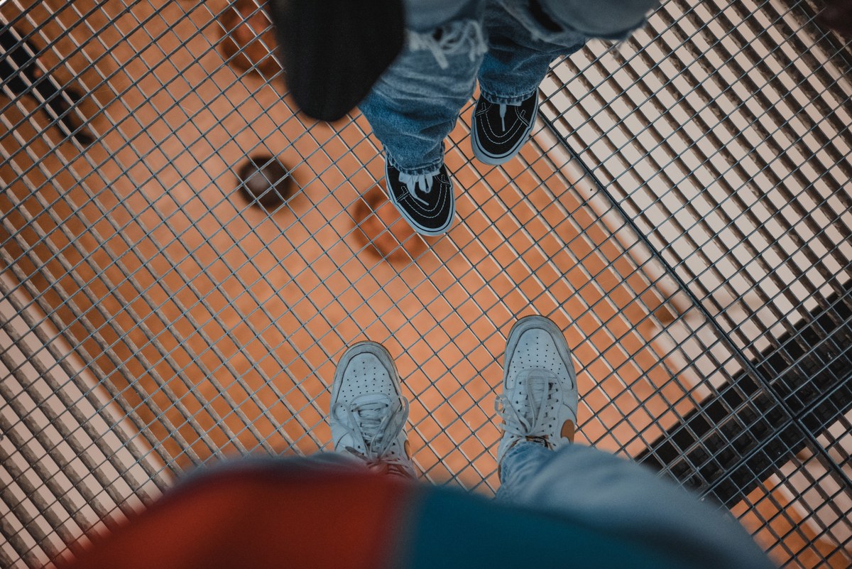 Twee mensen staan tegenover elkaar op een metalen rooster, gezien van bovenaf met hun sneakers zichtbaar boven een oranje ondergrond.