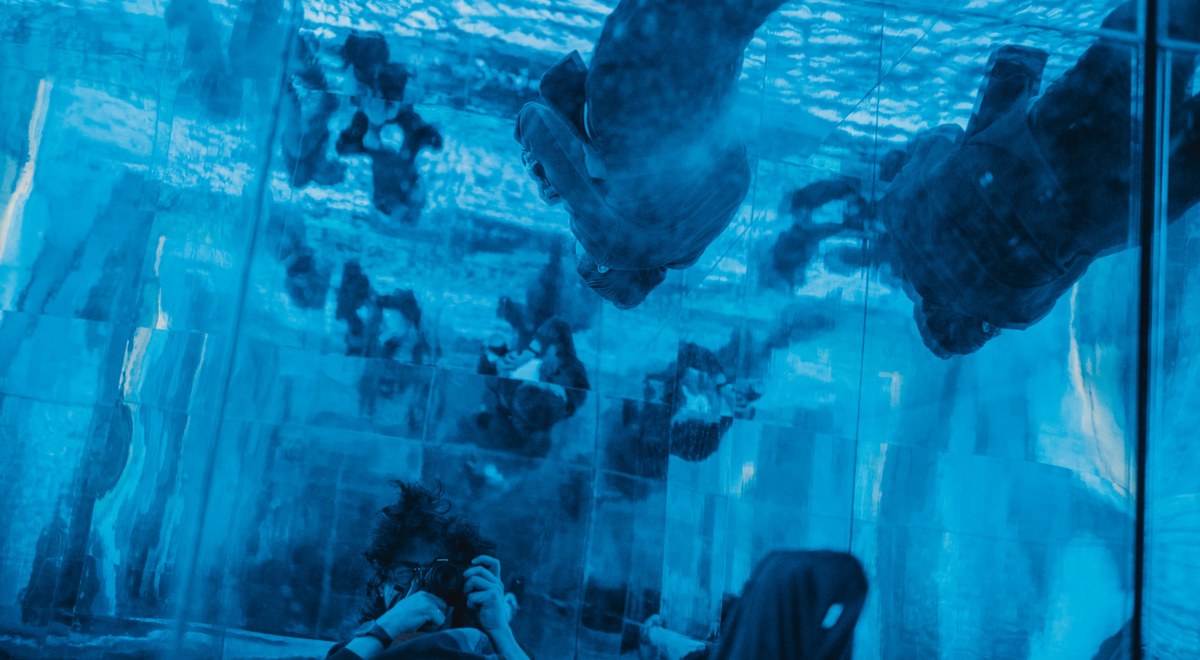 Bezoekers kijken van onderaf omhoog door een blauw verlicht wateroppervlak waarop mensen lijken te lopen, in een spiegelende installatie in de Kunsthal.