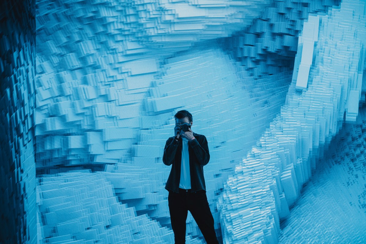 Een man in een zwart jasje maakt een foto met zijn camera, omringd door een golvende blauwe installatie van uitstekende blokjes.