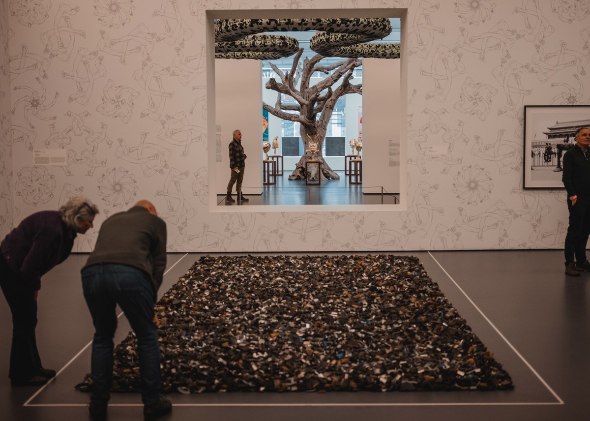 Twee bezoekers buigen zich over een groot vloerwerk van talloze kleine objecten, terwijl door een muuropening een boomsculptuur en meer kunst zichtbaar zijn.