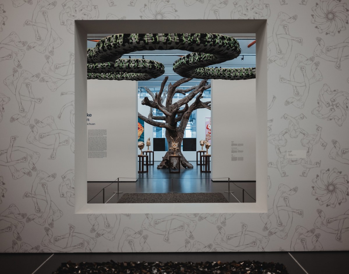 Door een vierkante muuropening met bloemenprint kijk je een museumzaal in, waar een grote boomsculptuur met groene bladerkronen centraal staat.