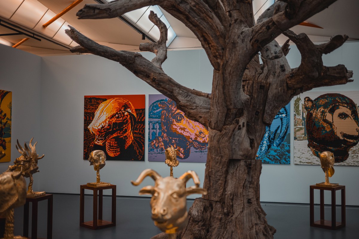 Een grote kale boom domineert een tentoonstellingsruimte met gouden dierenkoppen op sokkels en kleurrijke schilderijen van dieren aan de muren.