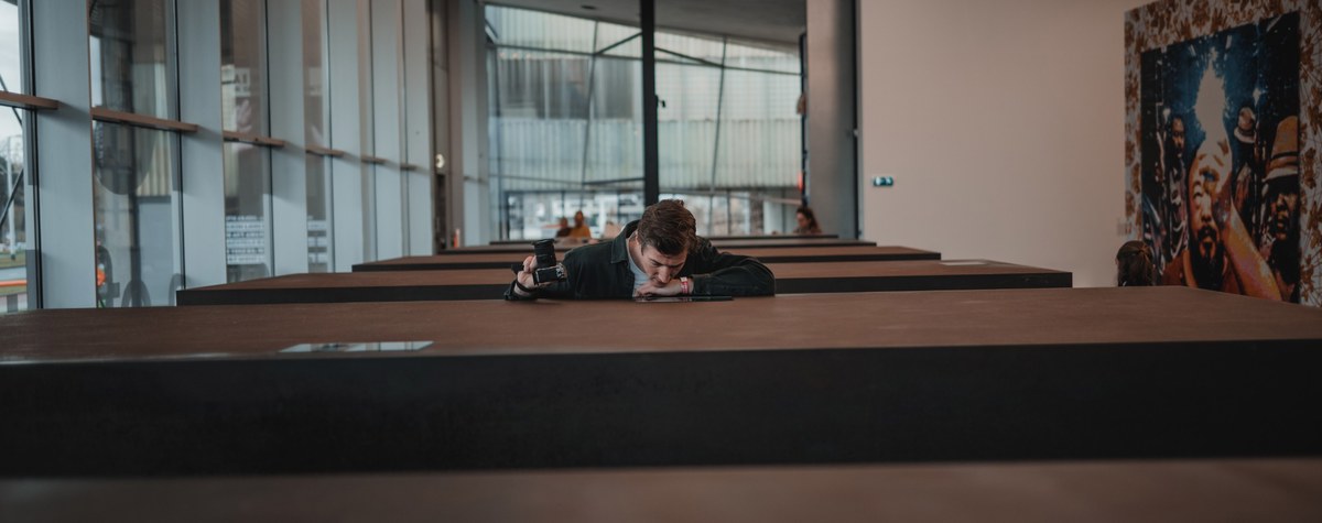 Een man leunt met zijn hoofd op zijn armen over een lange donkere bank in een moderne ruimte met grote ramen en een kleurrijk kunstwerk.