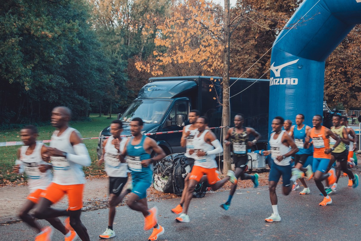 Een groep hardlopers in oranje en blauwe wedstrijdkleding passeert een zwart bestelbusje en een blauwe Mizuno-boog langs een herfstige bosrand.