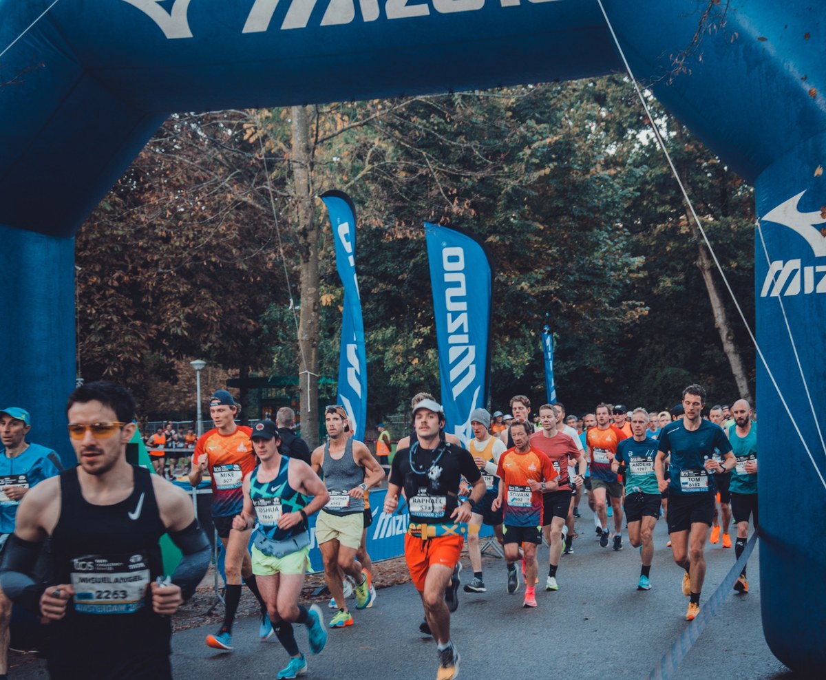 Hardlopers passeren de blauwe Mizuno-startboog tijdens de Amsterdam Marathon, met bomen en wapperende vlaggen op de achtergrond.