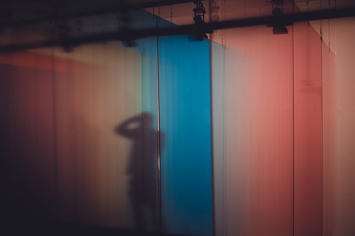 Silhouet van een persoon met opgeheven arm, geprojecteerd als schaduw op kleurige panelen in tinten roze, blauw en oranje.