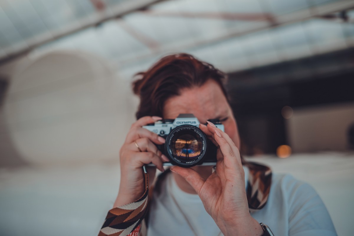Een persoon houdt een vintage Olympus-camera voor het gezicht en richt deze op de kijker, met een lichte, wazige industriële ruimte op de achtergrond.