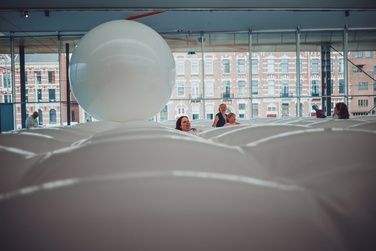 Bezoekers kijken tussen grote witte opblaasbare vormen door, met een grote witte bal en een bakstenen gebouw zichtbaar achter de glazen wand.